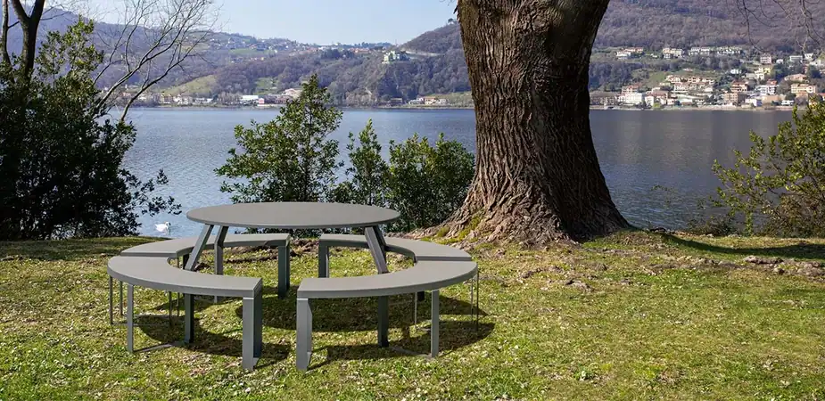 Sempreverde round picnic table with curved benches under a tree beside a lake